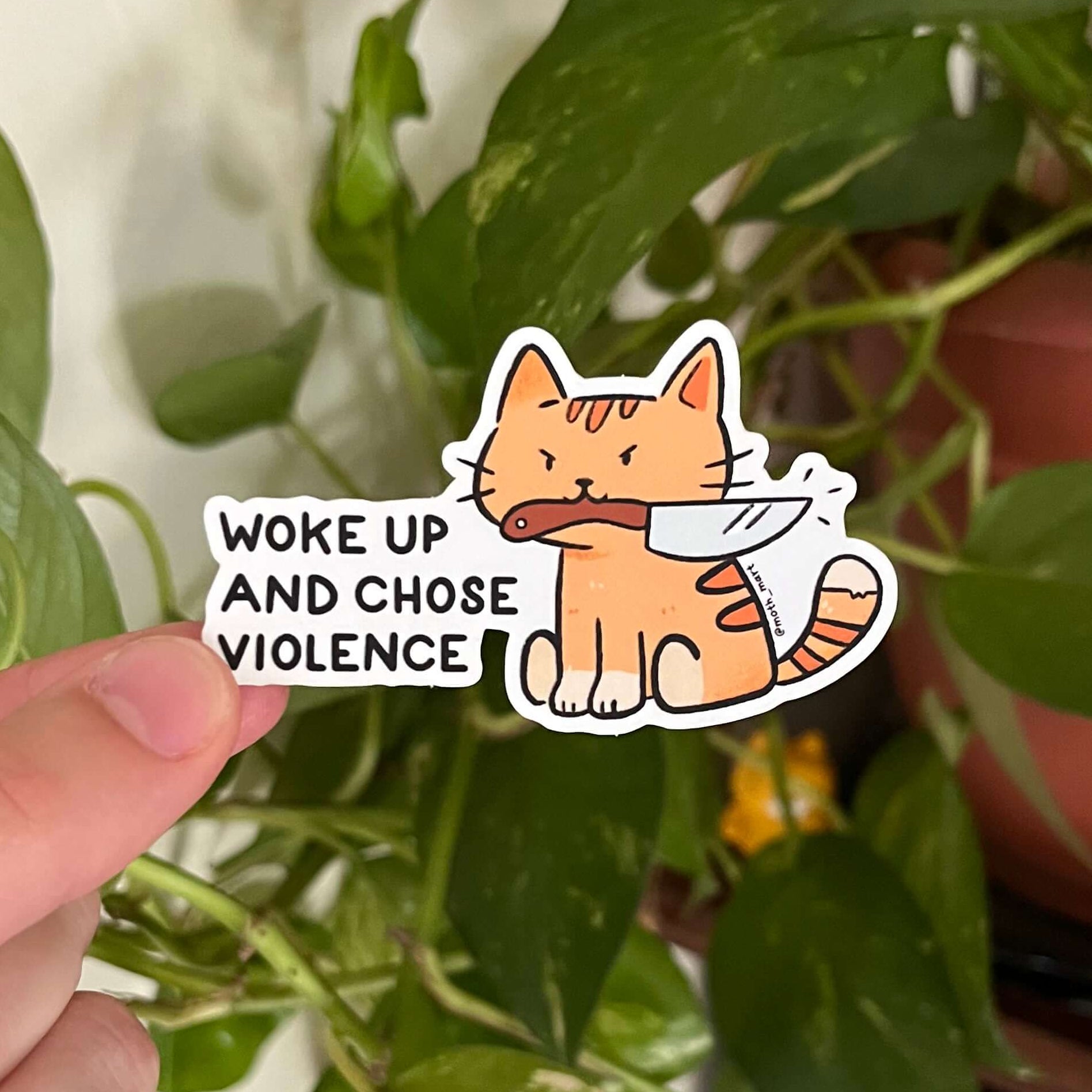 Sticker with a cat holding a knife and text 'Woke up and chose violence' held by a hand against a leafy background.