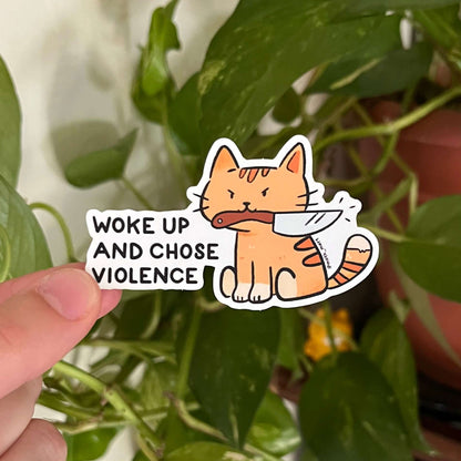 Sticker with a cat holding a knife and text 'Woke up and chose violence' held by a hand against a leafy background.