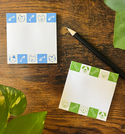 Two decorative sticky notepads on a wooden surface. The top notepad has blue cat and fish bone patterns, while the bottom one has green dog and bone designs. A pencil and plant leaves are nearby.