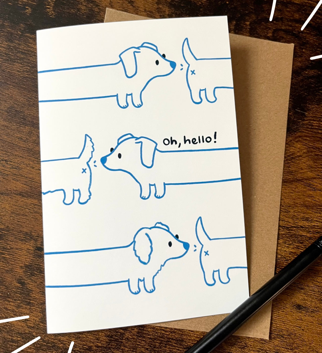 Greeting card on wood table with pencil of three dachshunds in blue line art, each sniffing the tail of the one ahead. The phrase "Oh, Hello!" is written playfully nearby. 
