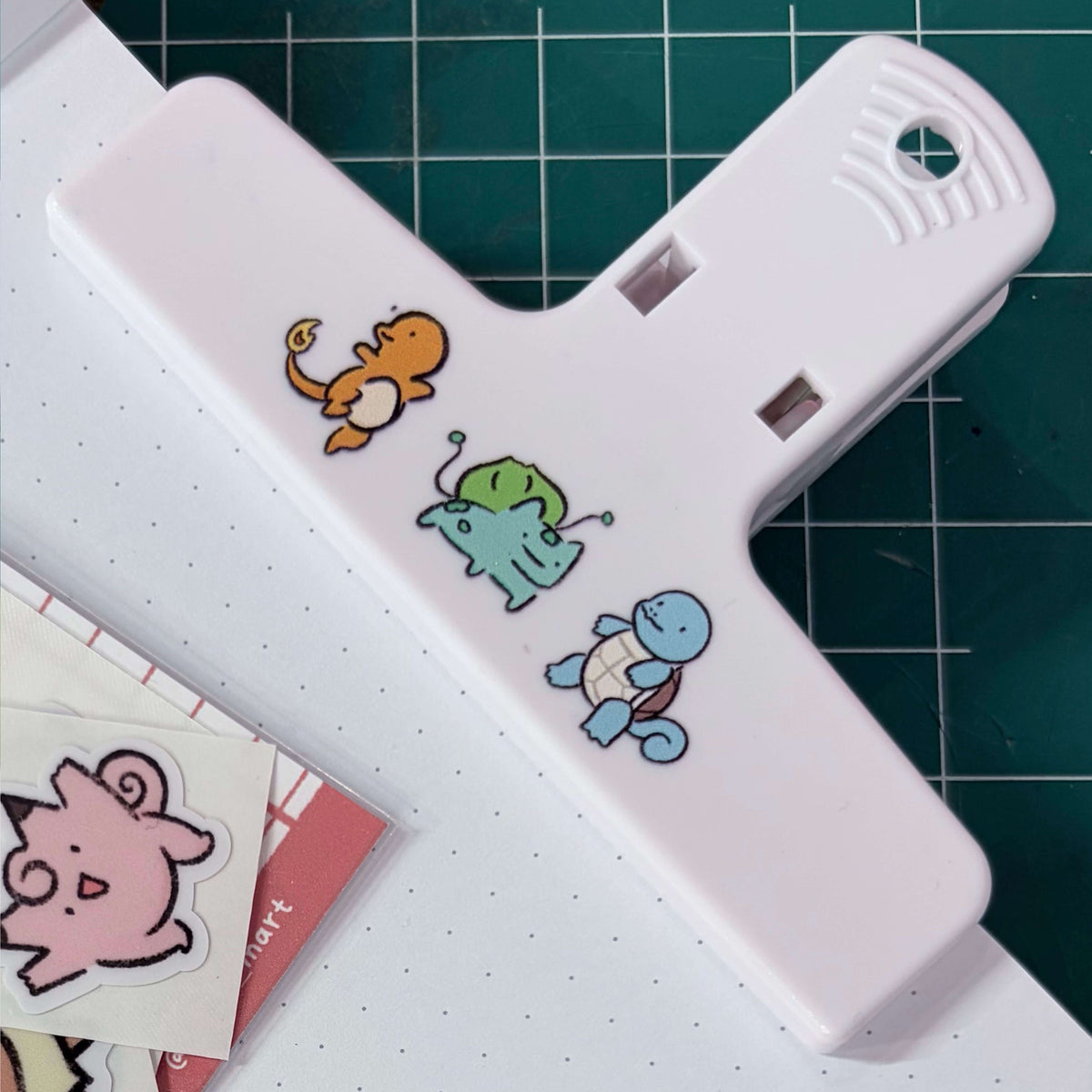 A white plastic clip or binder clip with three small cartoon characters on it, placed on top of a white dotted sheet over a green cutting mat.