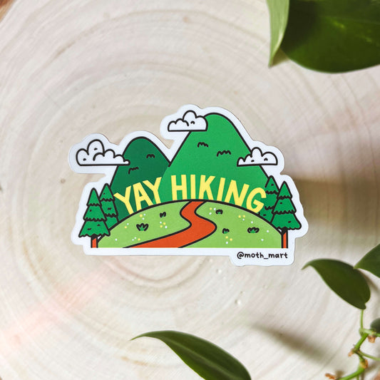 Sticker with 'Yay Hiking' design with hills and trees on a wooden surface with leaves.