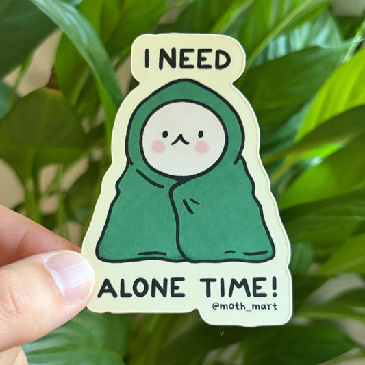 A die-cut sticker held by a hand against a background of green houseplants. The sticker features a cute cartoon character wrapped in a green blanket with the text 'I NEED ALONE TIME! @moth_mart'.