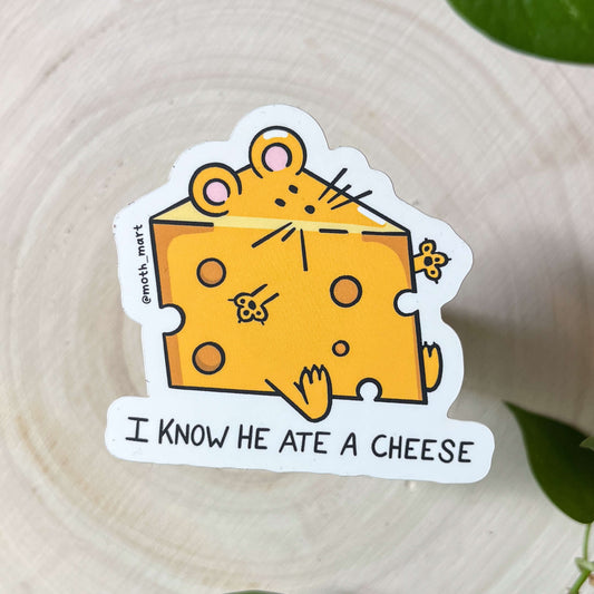 Sticker of a mouse inside a cheese wedge with text 'I KNOW HE ATE A CHEESE' on a light wood background