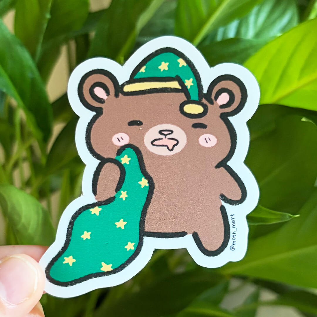 Die cut sticker of a sleepy brown bear wearing a green night cap with stars and holding a matching blanket. Held against a background of houseplants.