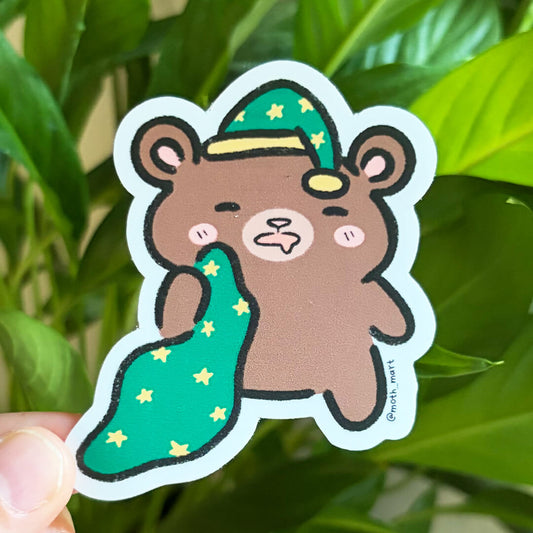 Die cut sticker of a sleepy brown bear wearing a green night cap with stars and holding a matching blanket. Held against a background of houseplants.