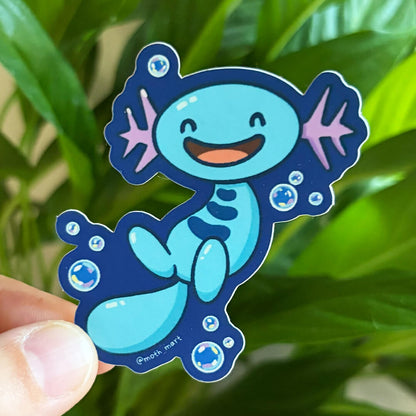 A die-cut sticker of a smiling blue axolotl with pink gills and bubbles, held by a person's fingers against a blurred green plant background.