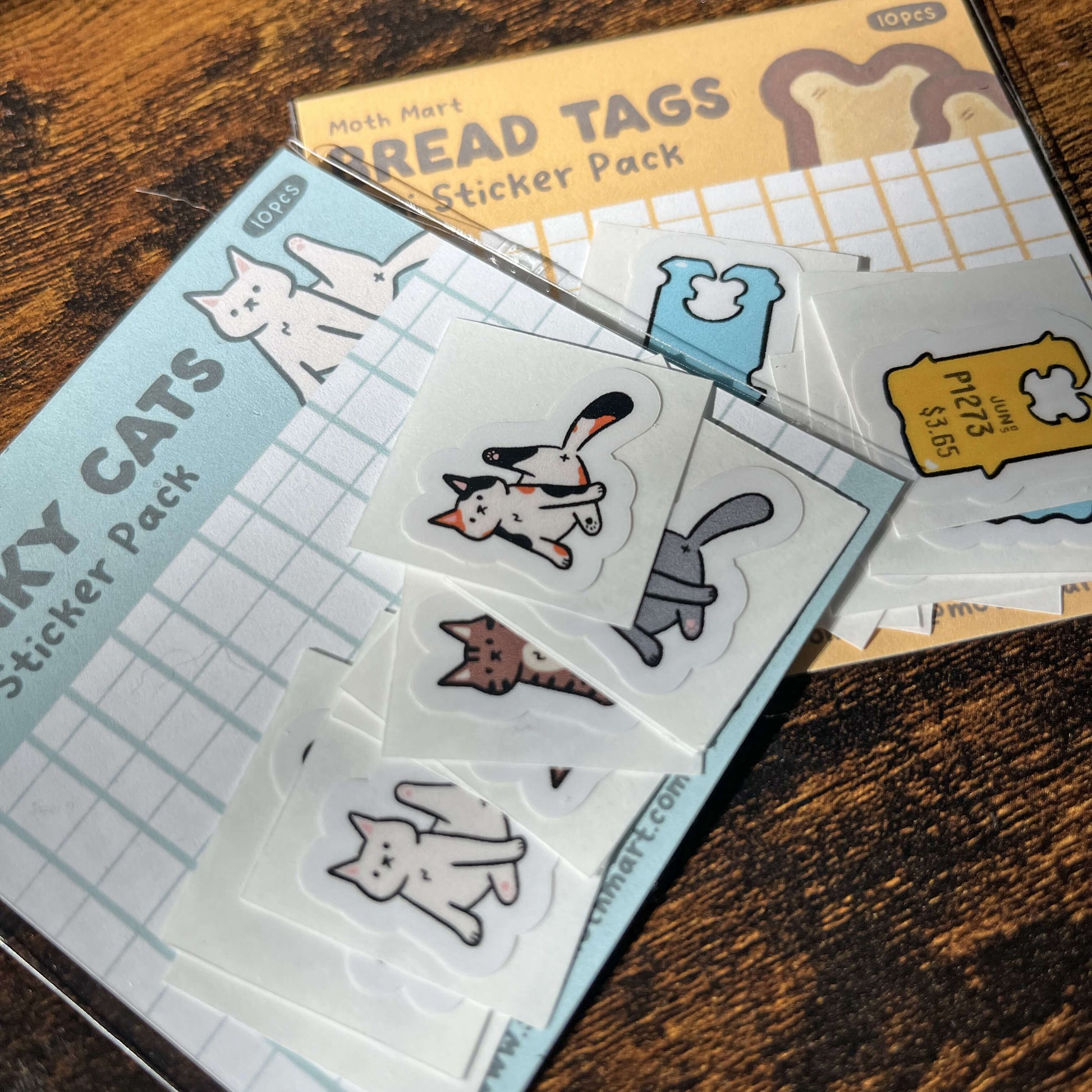Sticker packs with bread tags and cat designs on a wooden surface. Each come with 10 stickers.