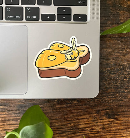 Sticker of a yellow moth laying on top of a piece of toast, melting like butter, on the corner of a laptop. 