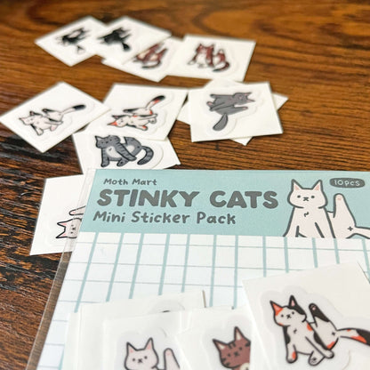 Stinky Cats mini sticker pack on a wooden surface. An additional 10 stinky cat stickers with their legs up are scattered around. 