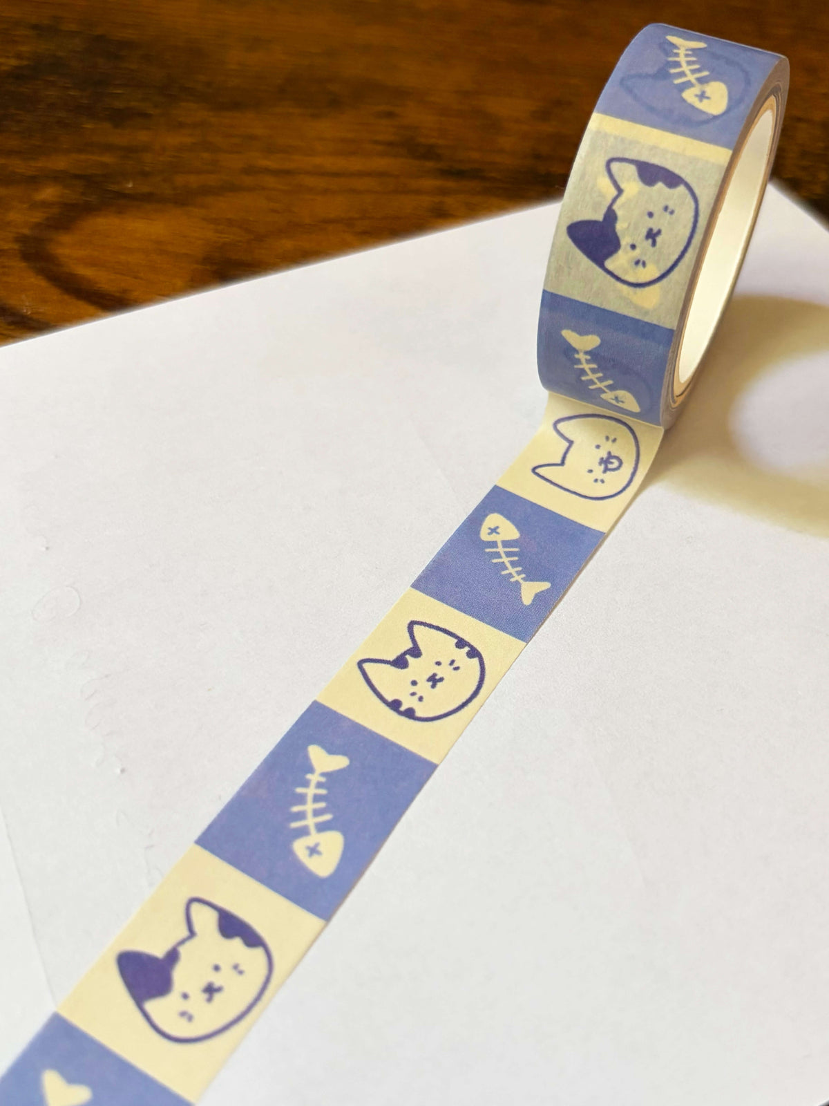 A roll of washi tape with a repeating pattern of blue cat faces and fish bones on a cream background, partially unrolled on a white surface with a wooden table in the background.