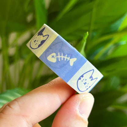 A hand holding washi tape featuring blue and cream cat illustrations and a fish skeleton motif, with a blurred green plant background.