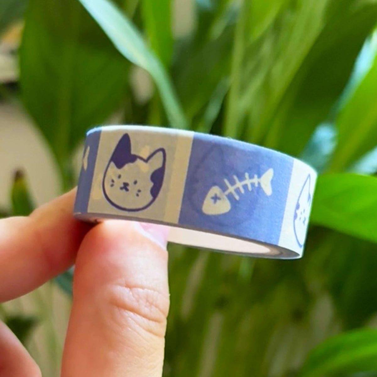 A hand holding a roll of blue and white washi tape with cat and fish bone illustrations, set against a blurred background of green houseplants.