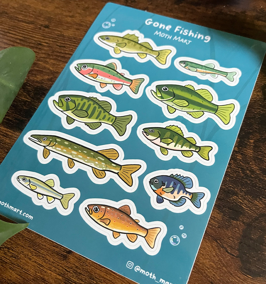 At an angle, stickers of various colorful freshwater fish on a blue background labeled "Gone Fishing." Wooden surface with green leaves.