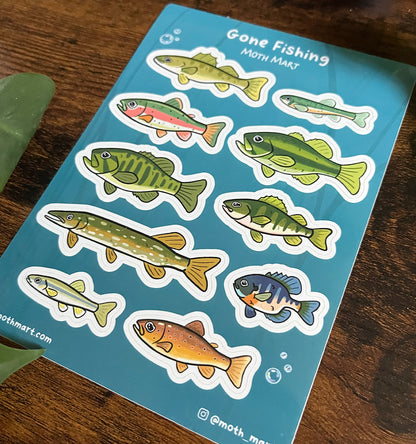 At an angle, stickers of various colorful freshwater fish on a blue background labeled "Gone Fishing." Wooden surface with green leaves.