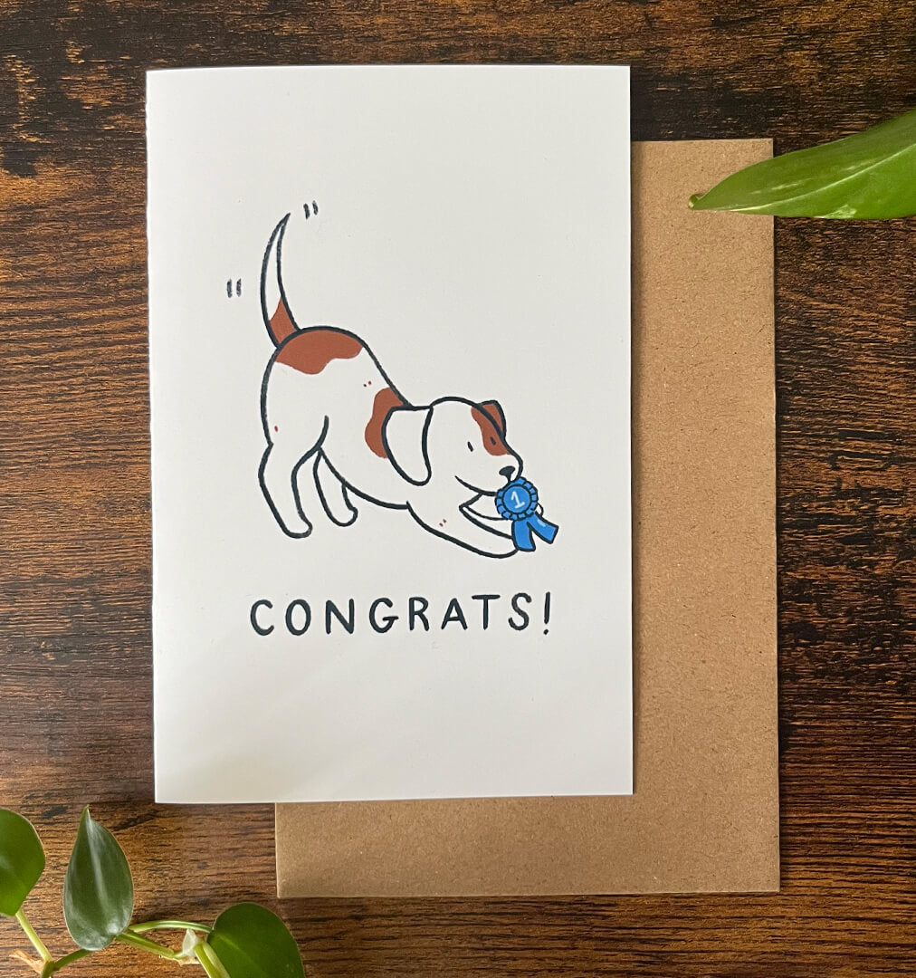 Greeting card with cartoon of a brown and white dog crouching and holding a #1 blue ribbon over "Congrats!" text on a wood background.
