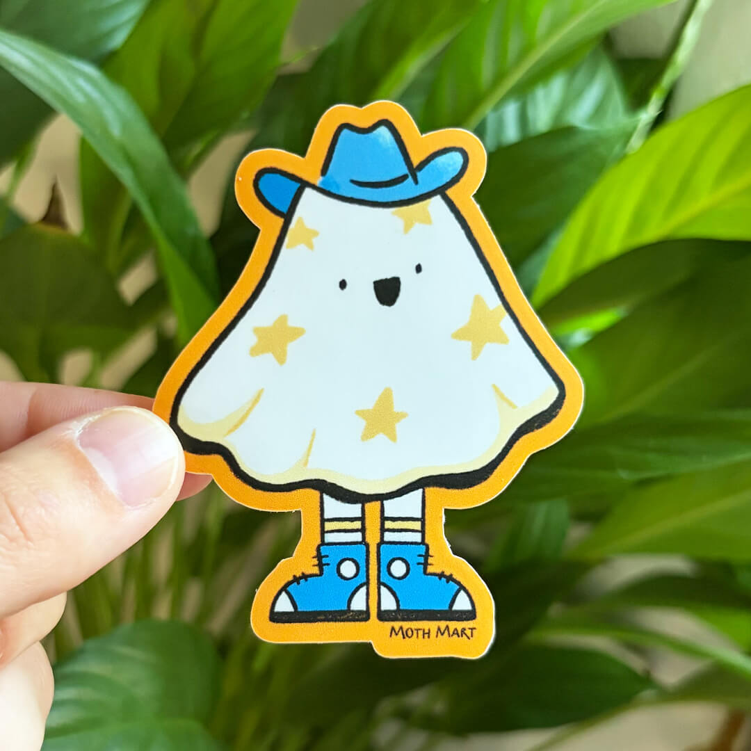 Die cut sticker of a ghost with stars on it, wearing a blue cowboy hat and blue sneakers with an orange background. Sticker is being held against a houseplant background. 