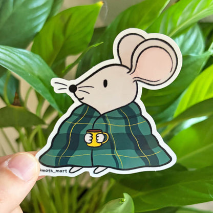 A die-cut sticker of a cartoon mouse wearing a green plaid blanket and holding a yellow mug, held by a hand in front of a green plant background.