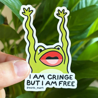 A die-cut sticker featuring a green cartoon frog with raised arms and the text 'I AM CRINGE BUT I AM FREE @moth_mart', held by a hand in front of a blurred green plant background.