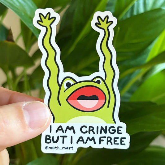 A die-cut sticker featuring a green cartoon frog with raised arms and the text 'I AM CRINGE BUT I AM FREE @moth_mart', held by a hand in front of a blurred green plant background.