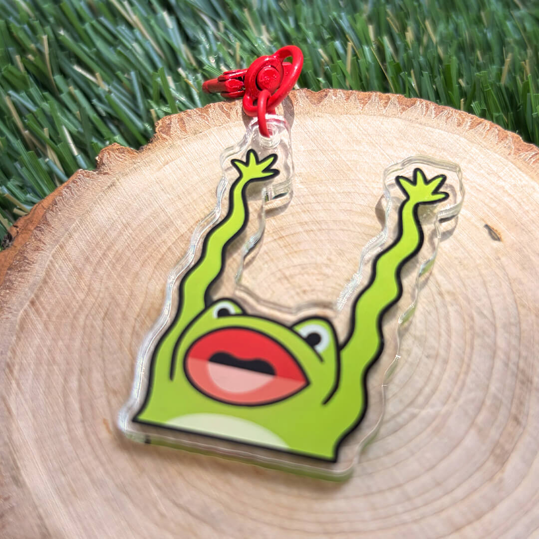 green frog keychain with its arms raised with red clasp on a wood disc on top of grass.