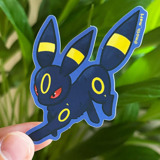 A die-cut sticker of a blue fox creature, held by a hand against a blurred background of green houseplants.