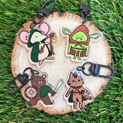 Four fantasy and gaming-themed animal character acrylic keychains displayed on natural wood slices, displayed on a green grass background.