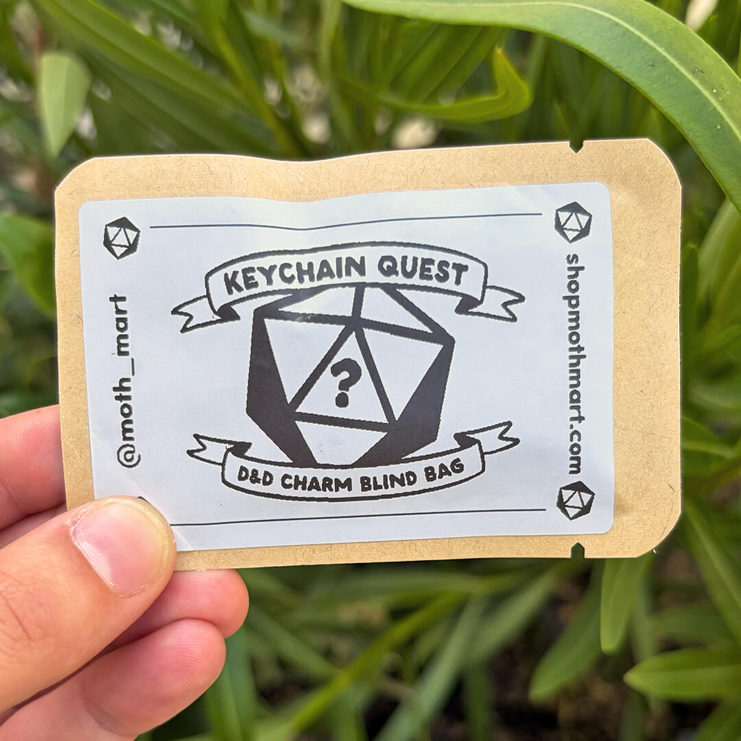 A hand holding a small kraft paper pouch labeled 'Keychain Quest D&D Charm Blind Bag' in front of green foliage. The pouch has a white label with black text and a large 20-sided die graphic.