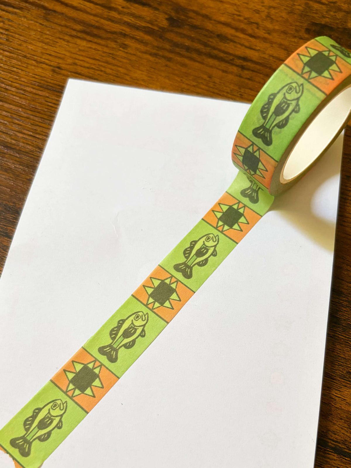 A roll of washi tape with a green fish and orange quilt pattern partially unrolled over a white sheet of paper on a wooden table.