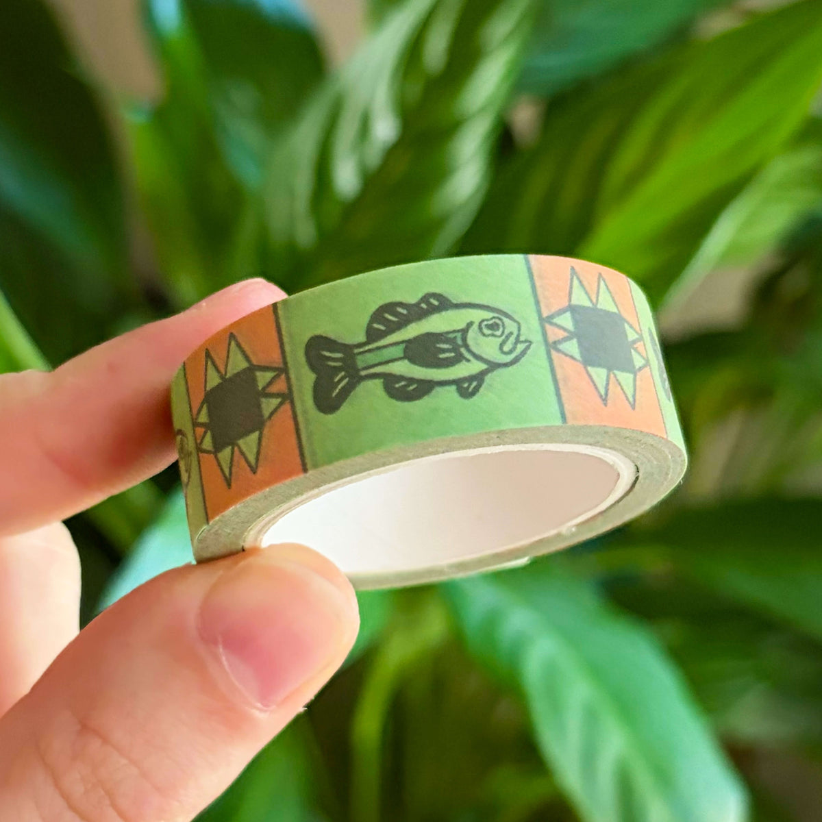 A hand holding a roll of decorative washi tape with a green fish and orange quilt pattern set against a blurred background of green houseplants.