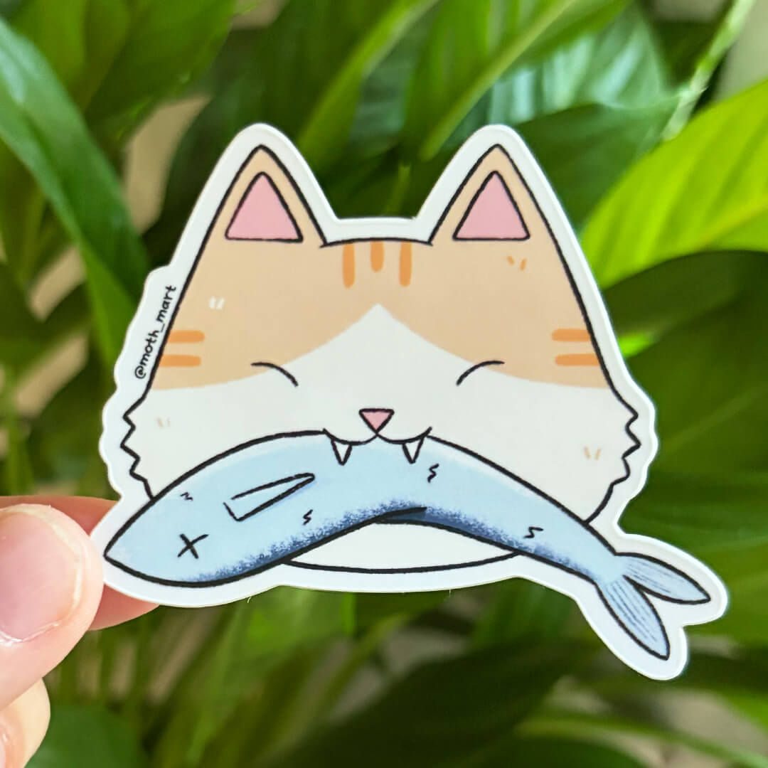 Sticker of an orange and white tabby cat head holding a blue fish, held by a hand in front of a leafy background. 