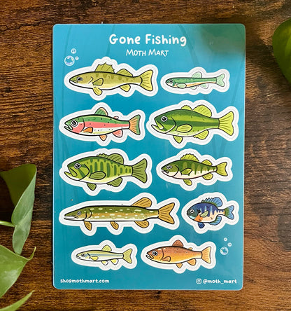 Stickers of various colorful freshwater fish on a blue background labeled "Gone Fishing." Wooden surface with green leaves surrounds the sheet.