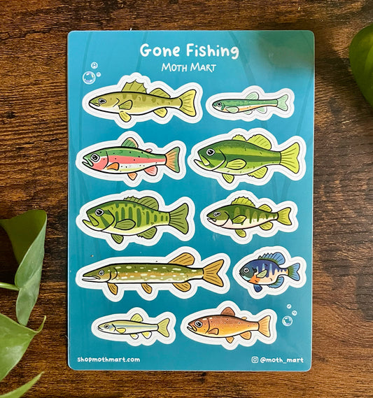 Stickers of various colorful freshwater fish on a blue background labeled "Gone Fishing." Wooden surface with green leaves surrounds the sheet.
