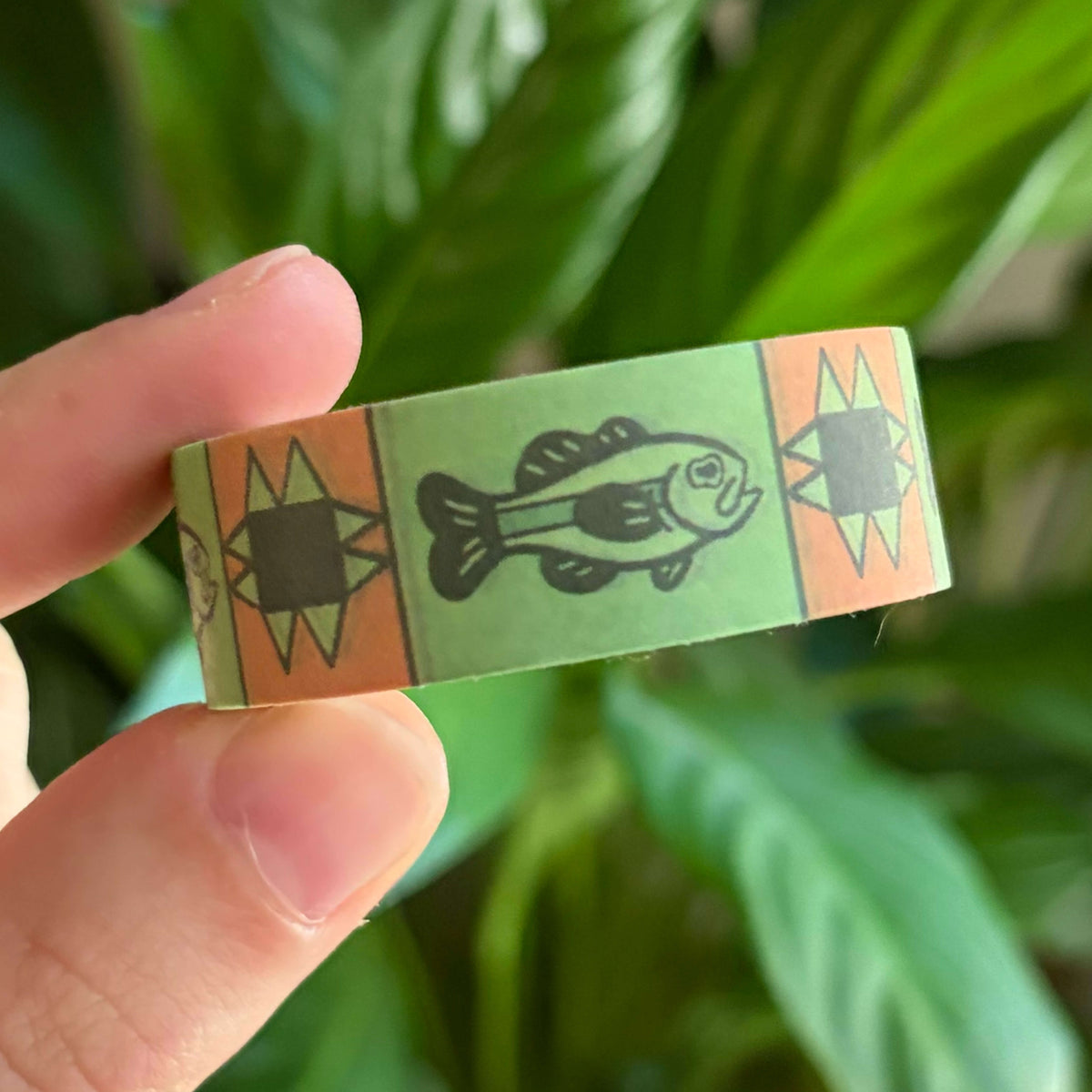 A close-up of a hand holding washi tape with fish illustration and orange geometric star motifs on a light green background, with blurred green leaves behind it.