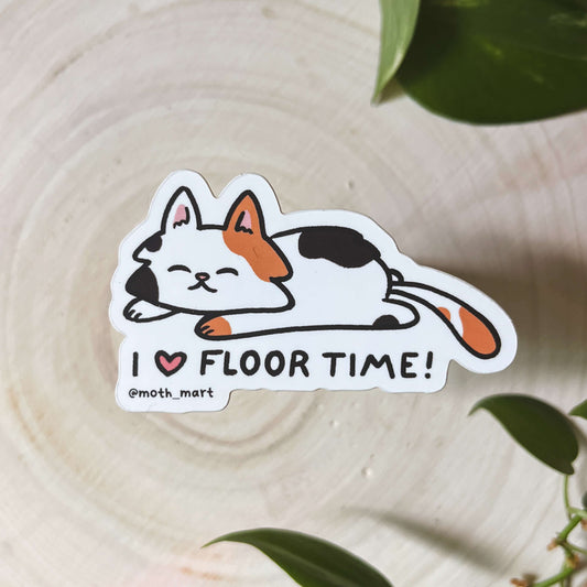 Illustration of a calico cat laying down with the text 'I Love Floor Time' on a wood background. 