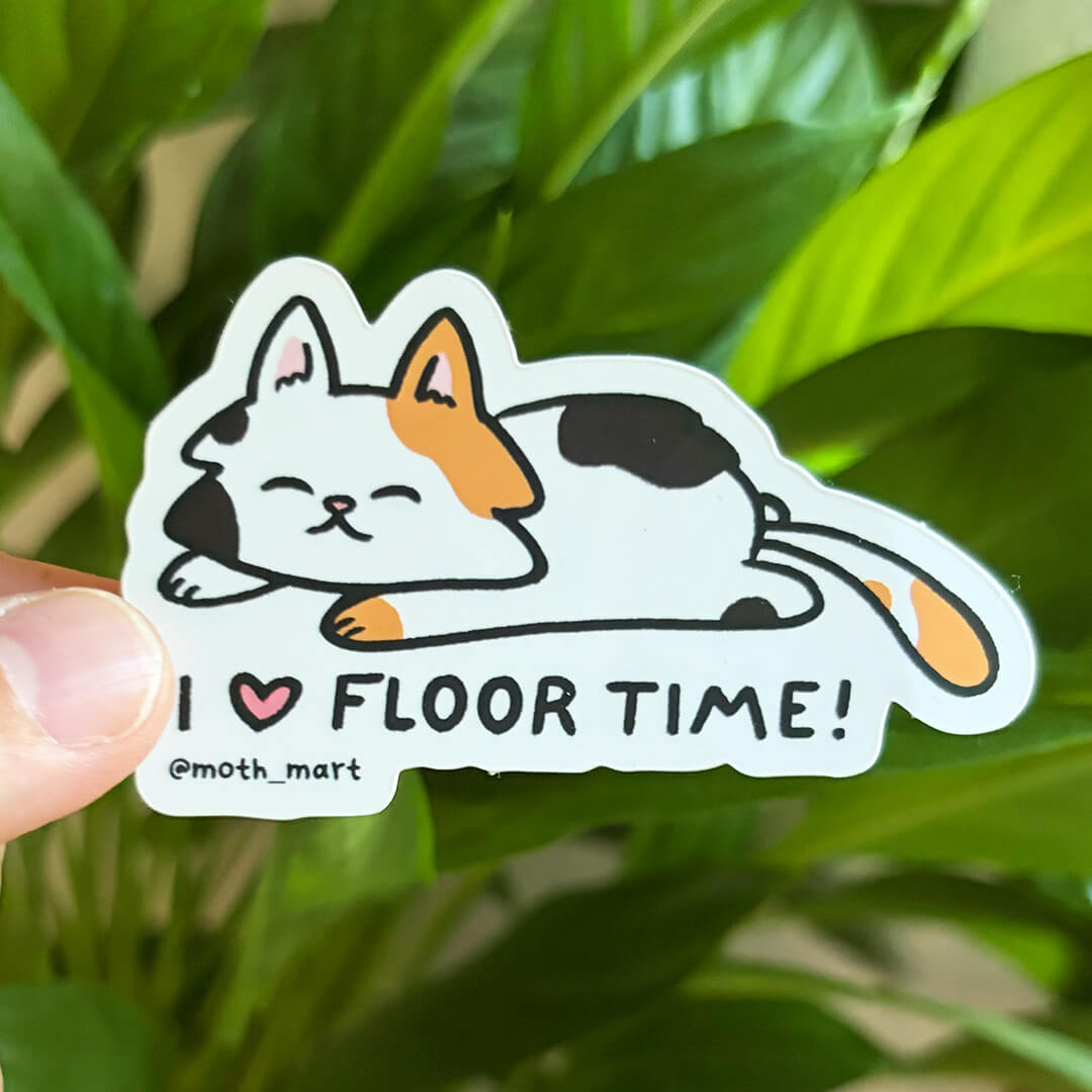 A die-cut sticker featuring a cartoon tri-color cat lying down with the text 'I [heart] FLOOR TIME!' and '@moth_mart', held by a hand in front of green houseplant leaves.
