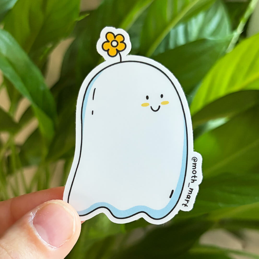 A hand holds a die-cut sticker featuring a cute white ghost character with a small yellow flower on its head, set against a blurred background of green houseplant leaves.