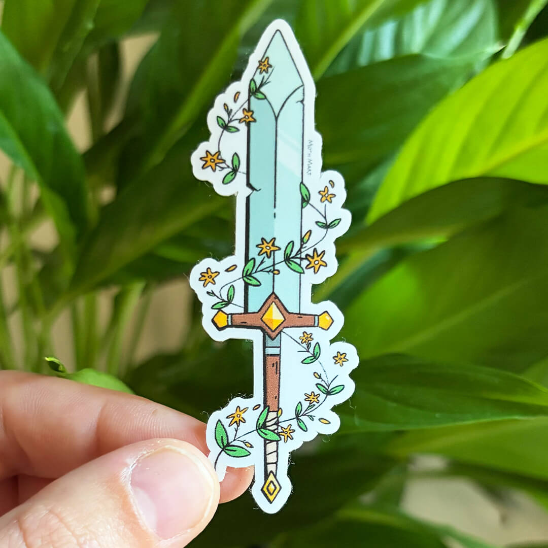 A die-cut sticker of a fantasy sword with yellow flowers and green vines, held by a hand against a background of green houseplant leaves.