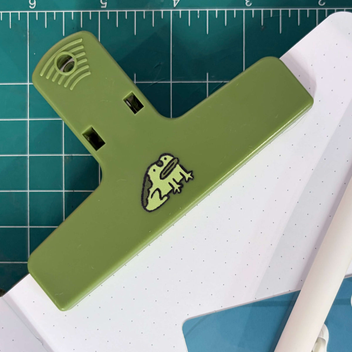An olive green T-shaped desk clip with a small cartoon frog design, shown holding a white dotted notebook on a green cutting mat with a white pen nearby.