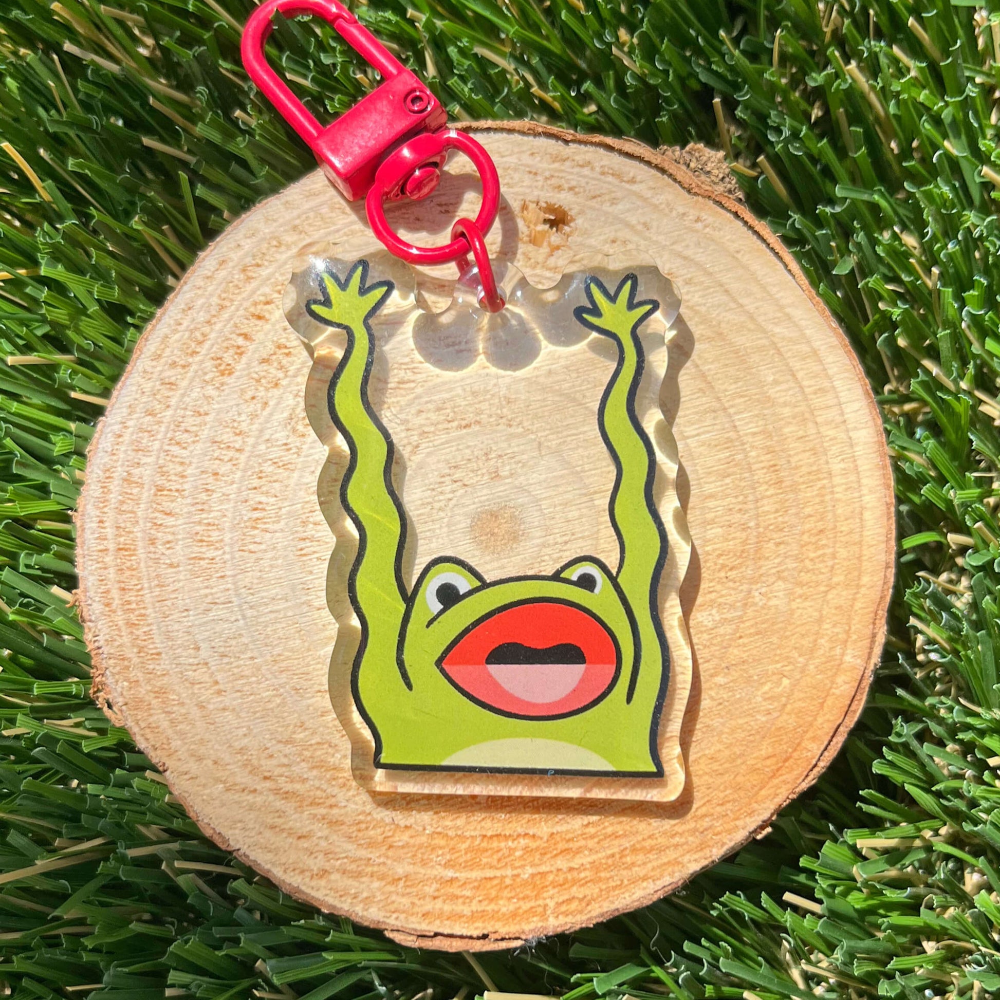 Keychain with a red hook and illustration of a frog, flailing its arms, on a wood coaster, placed on a grass background.