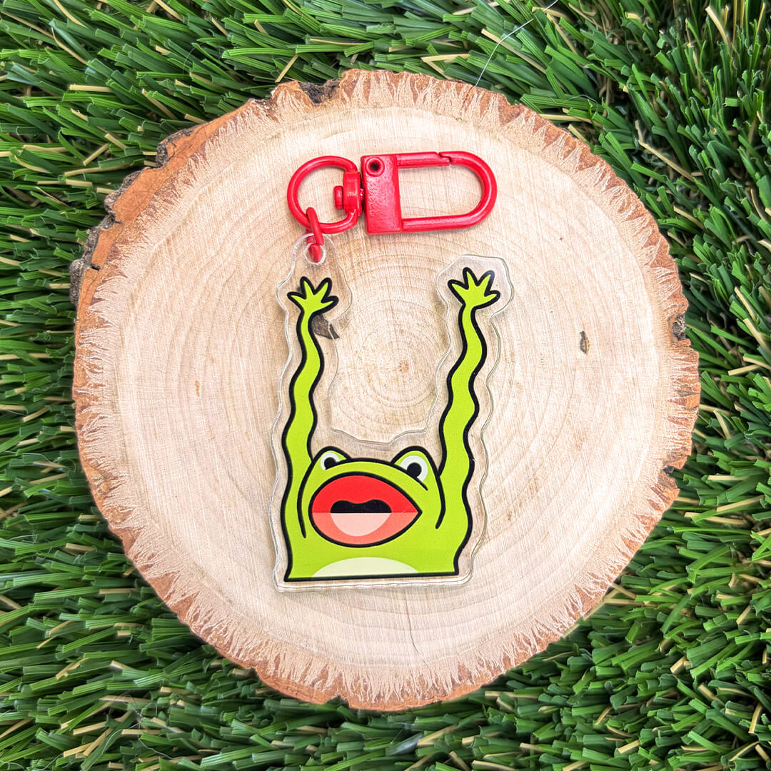 A green frog-shaped keychain with raised arms and a red carabiner, displayed on a round wood slice over green grass.