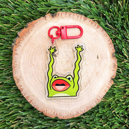 A green frog-shaped keychain with raised arms and a red carabiner, displayed on a round wood slice over green grass.