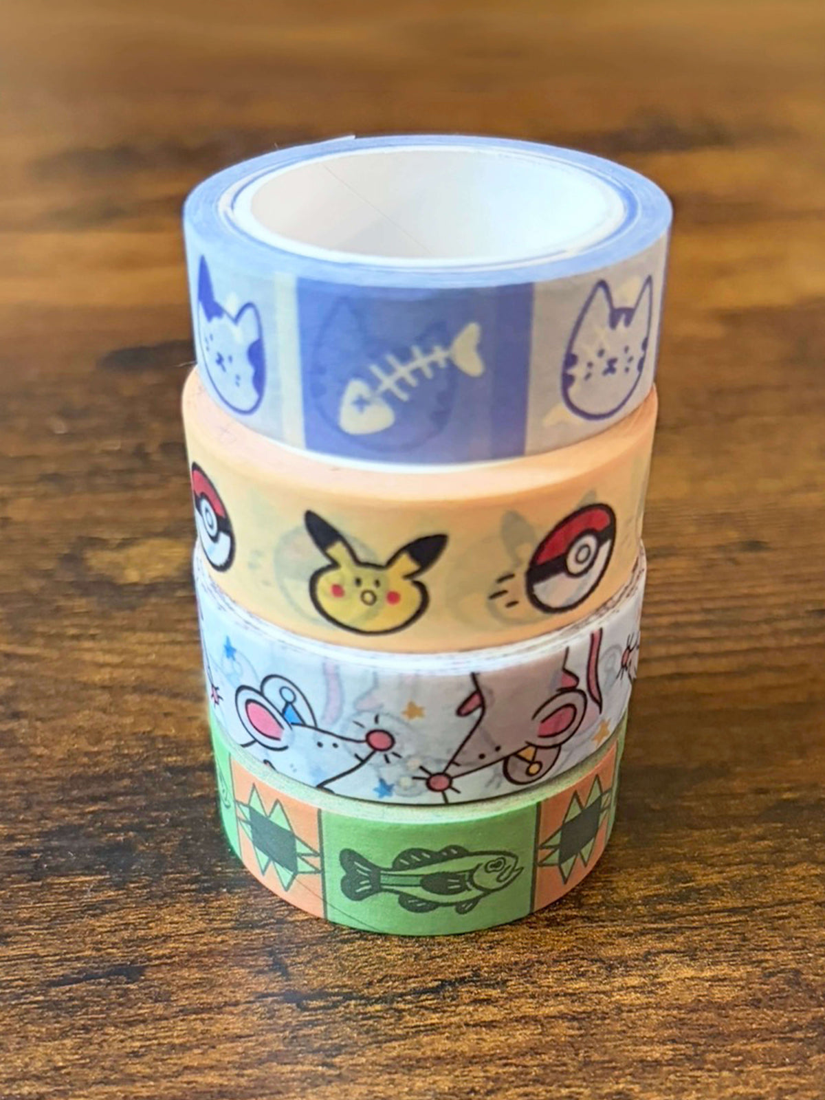 A stack of four rolls of washi tape featuring  rat designs, a cat skeleton, and a cat face design, a fish design, and a pocket critter design displayed on a wooden surface.