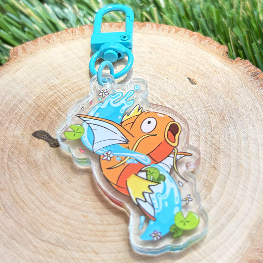 A clear acrylic keychain charm featuring a cartoon orange and yellow fish with blue water details, attached to a bright blue key ring and clasp, displayed on a round wooden slice with green grass in the background.