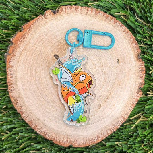 Orange koi fish acrylic keychain jumping out of water with a blue claw clasp on a wood disc, on top of grass