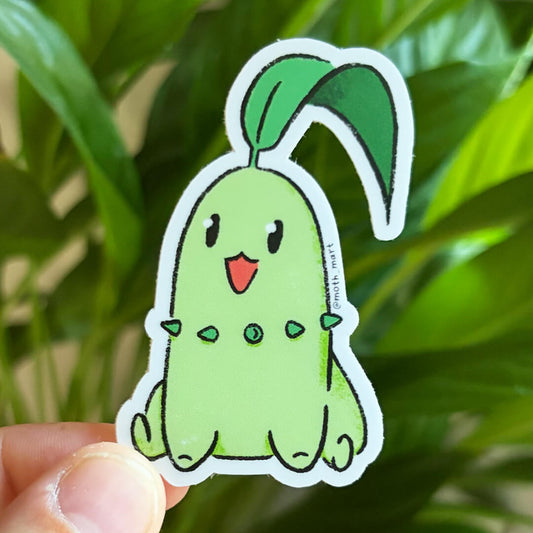 Die cut sticker of a round green critter with a leaf on its head held in front of a background of houseplants.