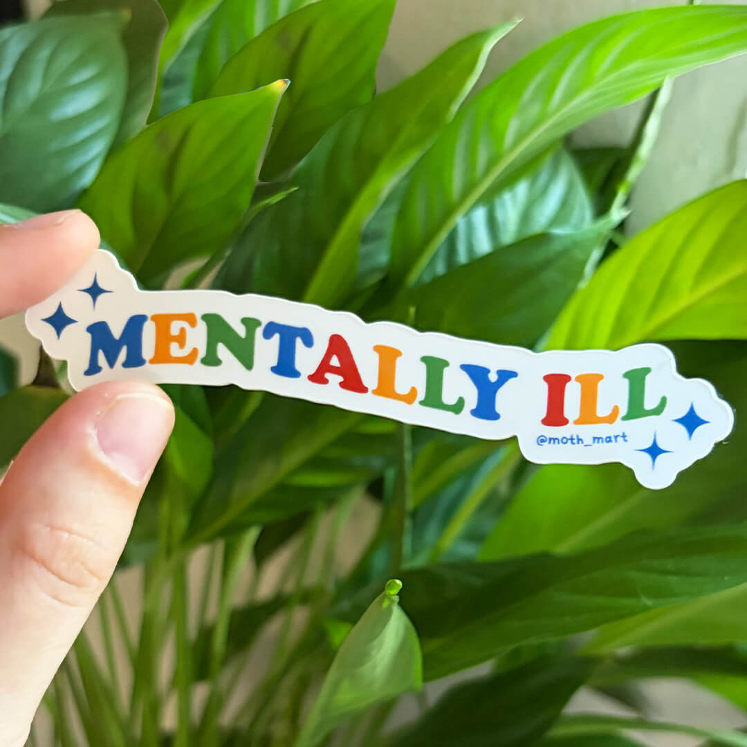 A hand holds a die-cut sticker that reads 'MENTALLY ILL' in colorful letters with blue star accents, set against a background of green houseplant leaves.