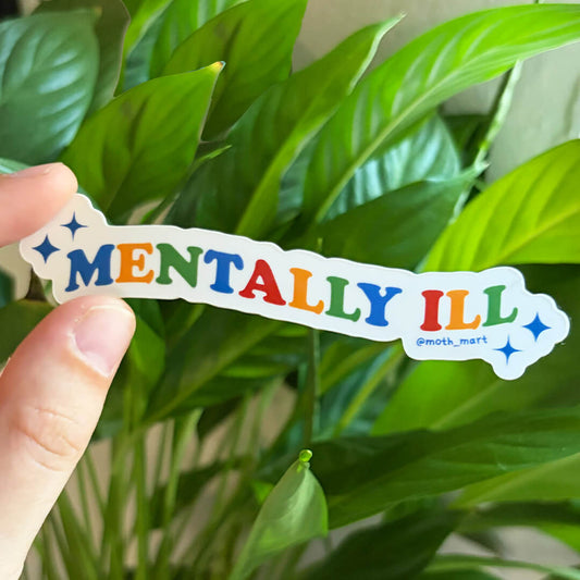 A hand holds a die-cut sticker that reads 'MENTALLY ILL' in colorful letters with blue star accents, set against a background of green houseplant leaves.