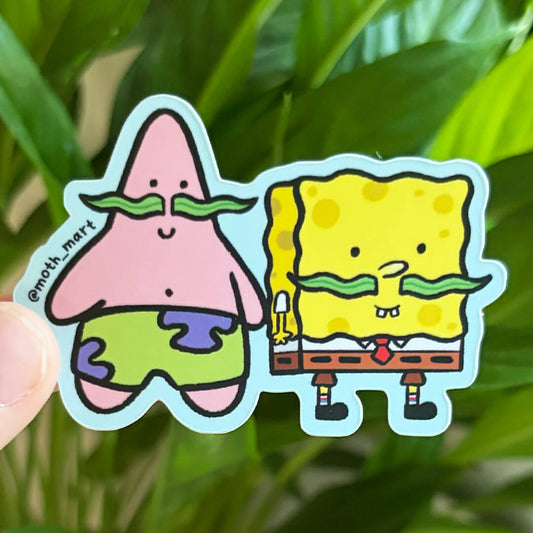 A close-up of a die-cut sticker featuring a sea star and sea sponge, both wearing green mustaches, held by a hand in front of a blurred green plant background.