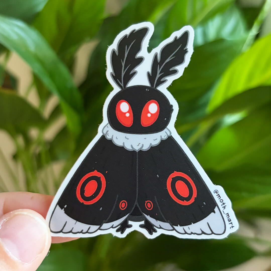 Dye cut sticker of a black and red mothman moth held against a background of house plants. 